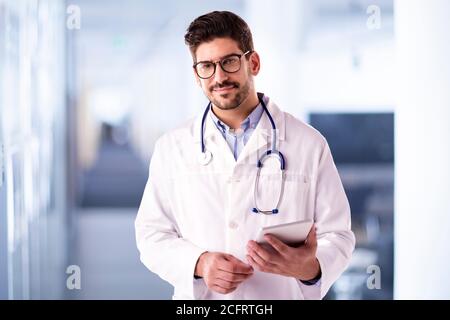 Aufnahme eines männlichen Arztes, der ein digitales Tablet in der Hand hält, während er im Foyer des Krankenhauses steht und arbeitet. Stockfoto