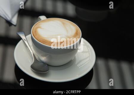Kleine weiße Tasse mit einem Design in der Kaffee-Schaum mit einem Teller, Löffel, Serviette, auf einer glatten schwarzen Oberfläche, wo Fensterbeleuchtung reflektiert wird Stockfoto