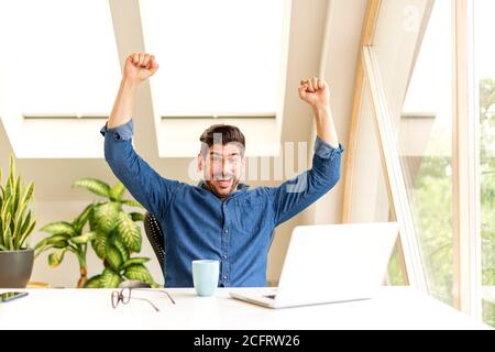 Fröhlicher Geschäftsmann feiert mit erhobenem Arm in der Luft, während er am Schreibtisch in seinem Heimbüro sitzt und an einem Laptop arbeitet. Heimbüro. Stockfoto