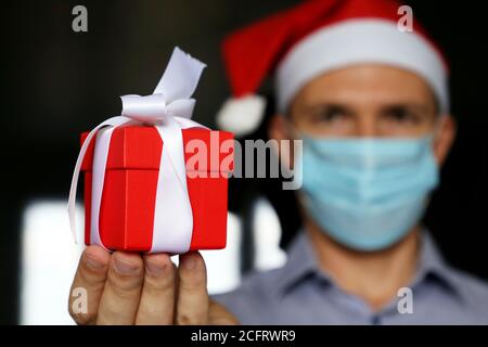 Rote Geschenkbox in der männlichen Hand Nahaufnahme, Mann in medizinischen Gesichtsmaske und Weihnachtsmann Hut geben Geschenk. Weihnachtsfeier während der Coronavirus-Pandemie Stockfoto
