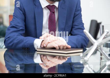 Nahaufnahme einer Person mit blauem Anzug, roter Krawatte und weißem Hemd, die an einem Glastisch sitzt und die Hände auf einem braunen Notizbuch zusammenfaltet Stockfoto