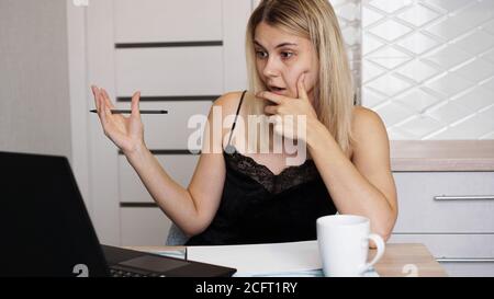 Porträt einer attraktiven Frau am Tisch mit Tasse und Laptop. Verwirrt junge Frau mit Problem mit Computer, Blick auf Laptop-Bildschirm Stockfoto