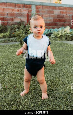 Liebenswert ein Jahr alten Jungen steht im Gras barfuß, hält sein Gleichgewicht, sieht verwirrt Stockfoto