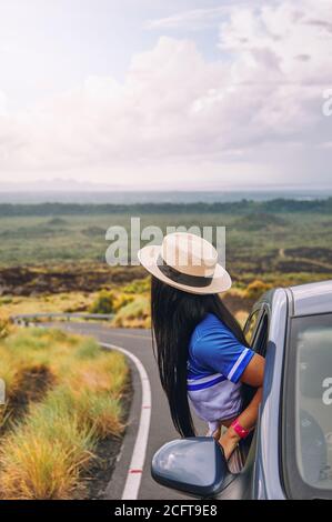 Eine junge Frau, die einen Hut trägt, der sich aus einem lehnt Autofenster mit einer schönen Landschaft im Hintergrund Stockfoto