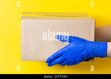 Ein Mann in medizinischen Latexhandschuhen liefert ein Paket auf gelbem Hintergrund. Das Konzept der sicheren Lieferung von Waren während der Coronavirus-Pandemie. Stockfoto