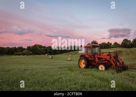 Roter Traktor und frisch gerollte Heuballen ruhen auf Rollen Hügel mit dramatischer Wolkenlandschaft bei Sonnenaufgang schaffen eine rustikale ländliche Szene Stockfoto