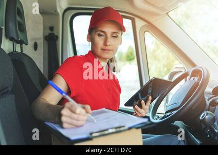Lieferperson in roter Uniform sitzt im Lieferwagen und schreibt Dokumente in der Zwischenablage Stockfoto