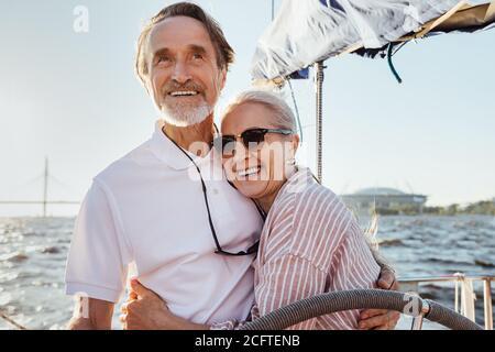 Fröhliches Seniorenpaar, das am Steuer auf einer Yacht steht und sich umarmt. Glückliche Menschen genießen sonnigen Tag auf einem Boot. Stockfoto