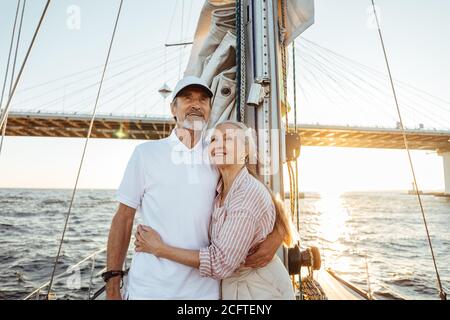 Glückliches Paar von zwei reifen Menschen genießen Urlaub auf privater Yacht. Lächelnde Senioren umarmen sich zusammen, während sie auf dem Segelbootdeck stehen. Stockfoto