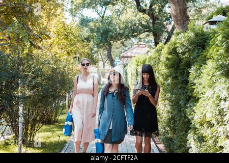 Drei schöne junge Frauen, die nach dem Einkaufen im Sommerpark spazieren gehen. Gruppe von internationalen Menschen. Stockfoto