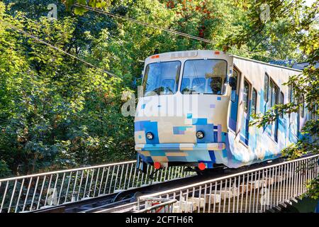 Eine blau-weiße Seilbahn, die von hellem Sonnenlicht beleuchtet wird, steigt auf Schienen entlang des Abhangs, umgeben von grünen Bäumen. Stockfoto