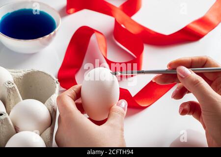 Junge Frau malt weiße Eier für Ostern. Eine junge Frau malt auf einem weißen Hintergrund Ostereier. Das Ei in der Hand des Mädchens Stockfoto