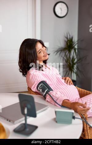 kaukasische schwangere Frau misst den Blutdruck mit automatischem Blutdruckmessgerät zu Hause, sitzen hinter dem Tisch in Pyjamas, am Morgen Stockfoto