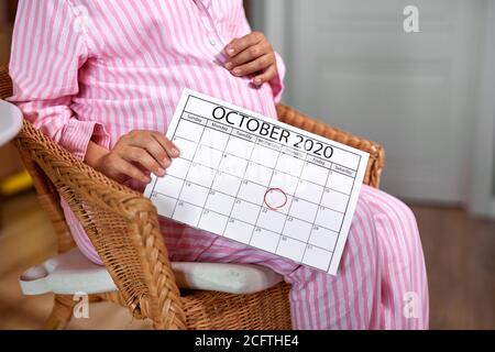 Schwangere Frau halten Kalender in den Händen, erwarten Baby. Zählen Tage, sie wird im oktober Geburt geben. Sitzen Sie auf Stuhl zu Hause tragen Pyjamas Stockfoto