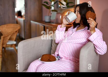 Schwangere Frau essen Schokoladenkekse und trinken Tee zu Hause, genießen Sie das Frühstück, leiden unter süßen Verlangen während der Schwangerschaft Konzept Stockfoto