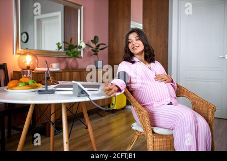 Glückliche kaukasische schwangere Frau Messung des Blutdrucks zu Hause, Gesundheits-Check der erwarteten Mutter. Frau lächeln, sitzt auf einem Stuhl neben dem Tisch Stockfoto