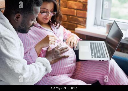 Schwarzer Mann ist freundlich zu seiner schwangeren kaukasischen Frau, junge männliche Schlaganfall Bauch, Frau im Pyjama sitzt mit Laptop zu Hause, verliebt Stockfoto