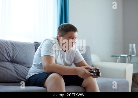 Keen kaukasischen Jungen spielen Videospiele, übergewichtigen Jungen sitzen auf dem Sofa allein, hat Probleme mit der Ernährung, führen ungesunden Lebensstil. Faulheit Konzept Stockfoto