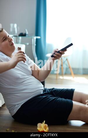 Fat Übergewicht Teenager Junge hat schlechte Ernährung, essen ungesunde Lebensmittel. Sitzen Sie auf dem Boden essen Junk-Food und Fernsehen Stockfoto