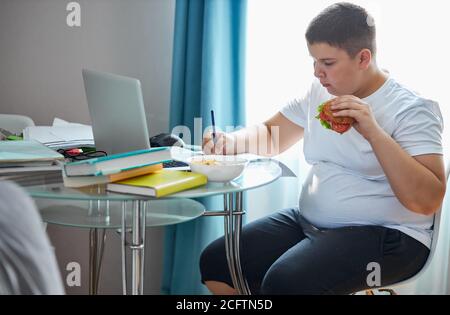 Übergewichtige Schuljunge essen Sandwich während Hausaufgaben, sitzt am Tisch konzentriert auf das Schreiben und halten Lebensmittel in den Händen Stockfoto