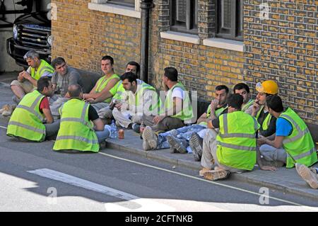 Londoner Straßenszene Gruppe von Baustellenarbeitern im Schatten auf dem Bürgersteig außerhalb der Baustelle in Warnjacken zur Mittagszeit in Großbritannien Stockfoto