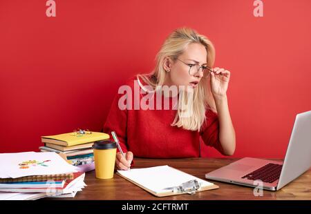 Junge Frau konzentriert sich auf die Arbeit am Laptop, sie schaut auf den Bildschirm und denkt, schreibt Informationen auf Papier Stockfoto