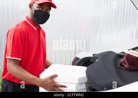 Selbstbewusster Deliveryman in roter Uniform legt Aufträge in das Auto, er wird es zu den Kunden tragen. Während der Quarantäne Stockfoto
