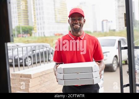 Lächelnder afro amerikanischer deliveryman mit Pizza in den Händen, gehen, um es für Kunden zu geben, Blick auf Kamera trägt rote Uniform Stockfoto