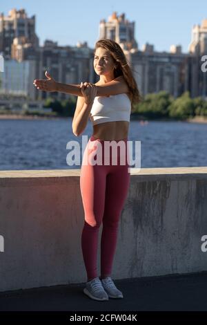 Fit junge sportliche Frau Strecken Arme und Schultergelenke auf Böschung. Fitness weiblich in rosa Leggings dabei Warmup Workout im Freien. Stockfoto