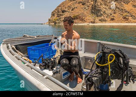 Ein Taucher sitzt auf dem Tenderboot der True North vor Komodo, Ost-Nusa-Tenggara, Indonesien, und bereitet ein Unterwasserkameragehäuse vor, mit Tauchflaschen in der Nähe. Stockfoto