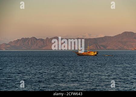 Ein traditioneller indonesischer Schoner glüht im goldenen Morgenlicht vor Anker vor Komodo Island, Indonesien, ein kleines Beiboot daneben festgemacht. Stockfoto