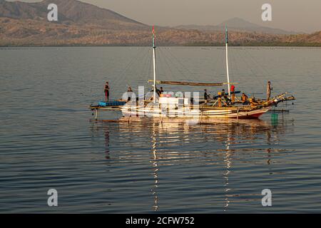 Ein Fischer steht am Bug eines Jukung-Außerbordboots vor der Küste von Lembata, Indonesien, während seine Kameraden Netze und Ausrüstung an Deck handhaben. Stockfoto