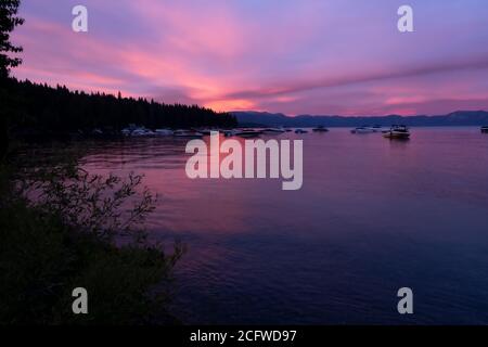 Strahlender Sonnenuntergang über Lake Tahoe, Laub und tiefes Wasser im Vordergrund, Boote, Wald, Berge im Hintergrund Stockfoto