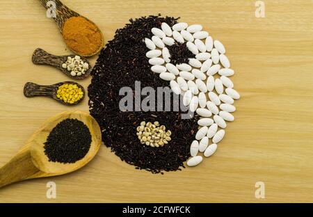 Schwarzer Reis und weiße Pille bilden ein Yin Yang Symbol und Kurkuma-Pulver, Hirse, Sojabohne, Basilikumsamen in Holzlöffel auf braunem Holz zeigen BL Stockfoto
