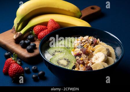 Obst, Müsli und Joghurt werden in einem Behälter vor dunkelblauem Hintergrund serviert. Eine Banane, Erdbeeren und Heidelbeeren daneben. Stockfoto