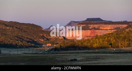 Eine leere Straße, die bei Sonnenaufgang in den Zion National Park führt Mit einem alten LKW im Vordergrund und der Schlucht Wände und Bäume durch das Sonnenlicht in beleuchtet Stockfoto