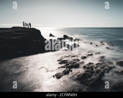 Eine lange Exposition von vier Menschen auf einer Klippe stehen Am Meer mit Felsen und Wellen im Vordergrund Und der Horizont und der Himmel im Hintergrund Stockfoto