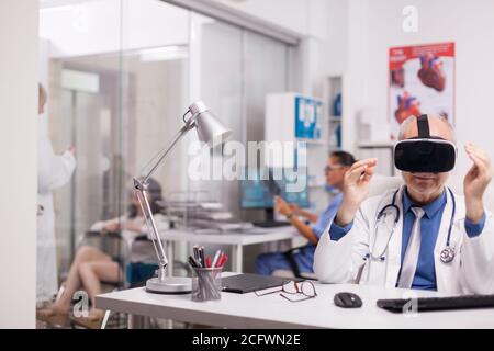 Älterer Arzt mit Virtual-Reality-Brille im Krankenhausbüro. Junge Therapeutin im Gespräch mit behinderten Frau im Rollstuhl auf Klinikkorridor und Krankenschwester hält Röntgen. Stockfoto