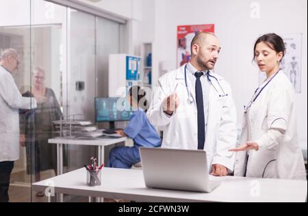 Team von Ärzten diskutieren Patienten Glauben Überprüfung diagnostizieren auf Laptop im Krankenhaus Büro. Senior Sanitäter und ältere Frau im Alter im Klinikkorridor und Krankenschwester in blauer Uniform arbeiten am Computer. Stockfoto