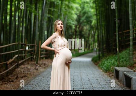 Schwangere Frau in Abendkleid hält Hände auf Bauch im Freien in Bambuswald. Schwangerschaft, Mutterschaft, Vorbereitung und Erwartung Konzept. Wunderschön Stockfoto