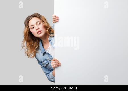 Junge Frau, die hinter einer weißen Wand herausguckt. Mockup der Ankündigung oder Präsentation. Stockfoto