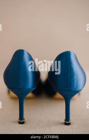 Verlobungsring der Braut mit einem Edelstein zwischen blauen Schuhen mit hohem Absatz auf dem Boden auf beigem Hintergrund. Stockfoto