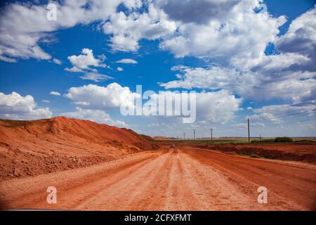 Bauxit-Tonerde-Tagebau. Rote Lehmstraße in Aluminiumerz-Steinbruch. Haufen von leeren Steinen und Gras vor blauem Himmel mit Wolken Hintergrund. Stockfoto