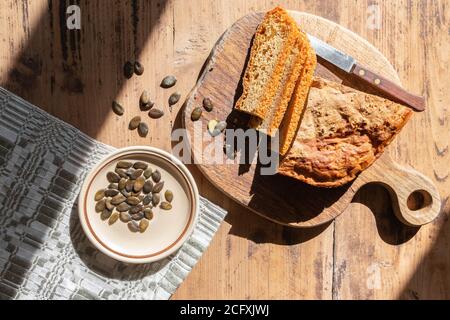 Hausgemachtes gebackenes Kürbisbrot Brot mit Messer auf Schneidebrett und Teller mit Samen auf rustikalem Tisch. Hausgemachte Brotscheiben. Stockfoto