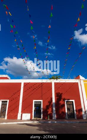 Lebhafte Fahnenketten über hellblauem Himmel mit weißen Wolken entlang rotem Kolonialgebäude, Valladolid, Yucatan, Mexiko Stockfoto