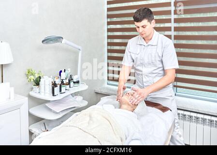 Männliche Kosmetikerin spendet Feuchtigkeit und reinigt das Gesicht der Frau mit Sahne und Seife. Spa-Behandlungen Stockfoto