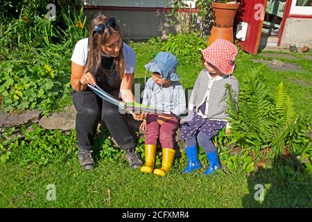 Frau trägt Latzhose Lesung ein Shirley Hughes Alfie Buch zu Junge und Mädchen Kinder im Alter von 3 sitzen außerhalb Haus in Rural Wales Großbritannien KATHY DEWITT Stockfoto