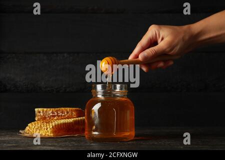 Weibliche Hand hält Dipper mit Honig auf Holzhintergrund mit Glas und Waben Stockfoto