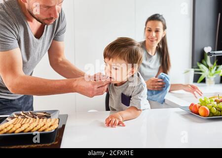 Vater gibt Kleinkind einen Keks zu Snack auf in der Küche mit Mutter im Hintergrund Stockfoto