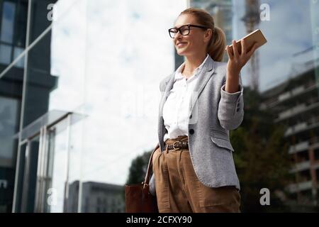 Glückliche erfolgreiche Geschäftsfrau trägt Brillen und klassische tragen halten Handy und lächelnd, während Sie im Freien gegen das Bürogebäude stehen Stockfoto